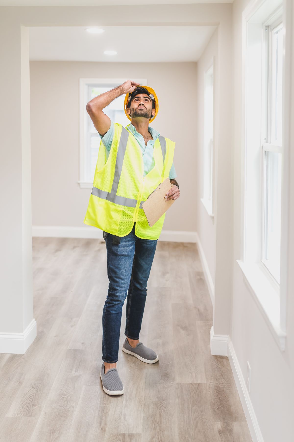 Inspector in a hard hat and safety vest standing in an empty room, looking up at the ceiling with a clipboard in hand