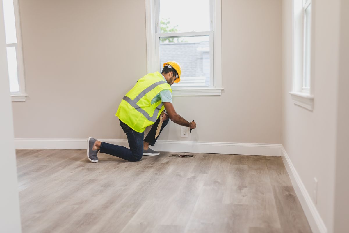 Inspector in a safety vest kneeling by a wall, checking an electrical outlet near the baseboard of an empty room